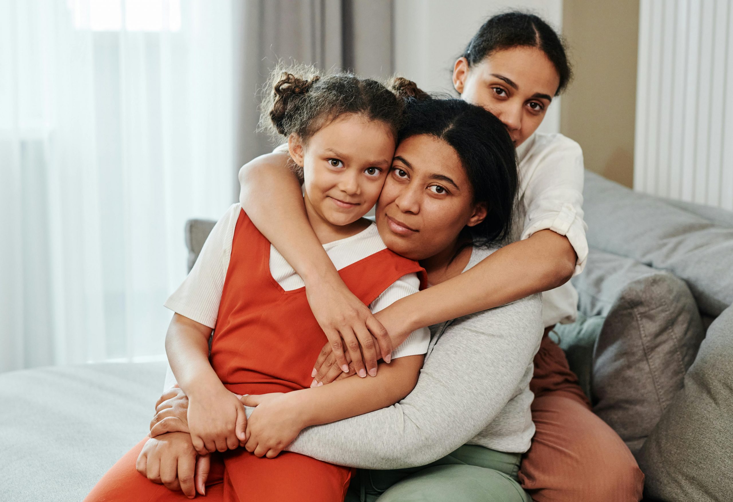 Young girl in orange dress seated between two women as they hug on a living-room couch, all looking at the camera.