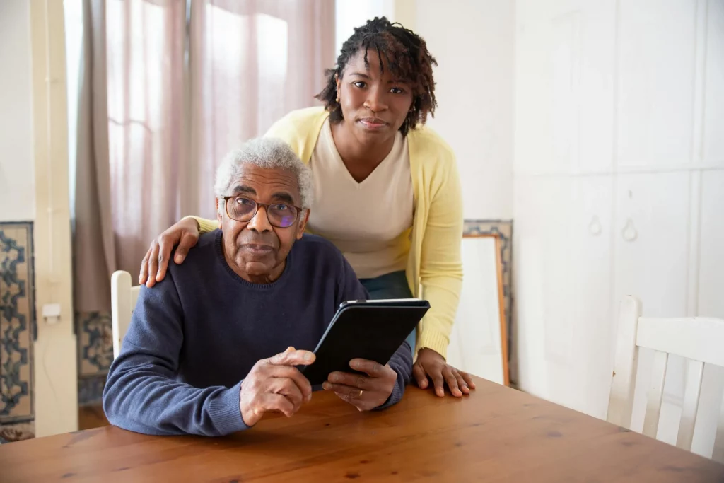 An older man sits at a table holding a tablet while a younger woman stands beside him with her hand on his shoulder.